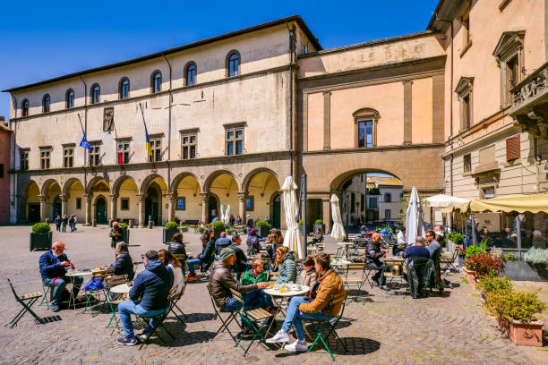 Customers and tourists enjoy life in Piazza del Plebiscito in the medieval heart of Viterbo Viterbo, Italy, April 18 -- Customers and tourists enjoy life in Piazza del Plebiscito in the historic heart of the medieval city of Viterbo, in central Italy. In the background, the Palazzo dei Priori, built in 1264 and also used as the seat of the Pontifical Government. In 1460 the building took on its current Renaissance style structure and is currently the Town Hall of Viterbo. The medieval center of Viterbo, the largest in Europe with countless historic buildings, churches and villages, stands on the route of the ancient Via Francigena (French Route) which in medieval times connected the regions of France to Rome, up to the commercial ports of Puglia, in southern Italy, to reach the Holy Land through the Mediterranean. Founded in the Etruscan era and recognized as a city in the year 852, Viterbo is characterized by its peperino stone and tuff constructions, materials abundantly present in this region of central Italy. Super wide angle image in high definition format. piazza plebiscito stock pictures, royalty-free photos & images