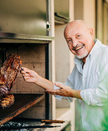 Portrait of smiling senior man grilling meat. Elderly man showing cooked meat on barbecue grill in backyard.
