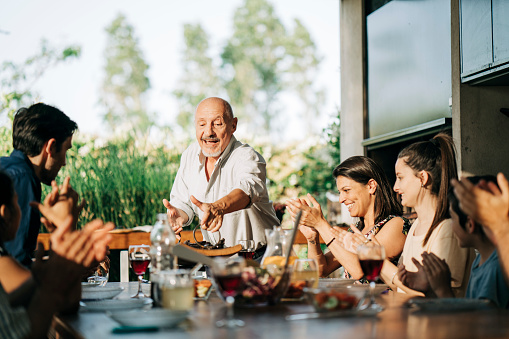 Cheerful senior man placing barbecue meat on tray over dining table for family. Multi-generational family at dining table in patio on a weekend.