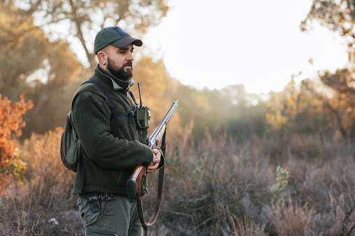Side view of young adult man with green clothes and cap holding his shotgun in middle of nature looking down with copy space.