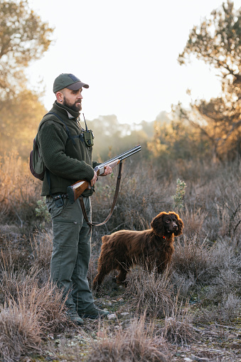 Side view of young hunter in green clothing standing and holding his shotgun in nature with his dog next to him.