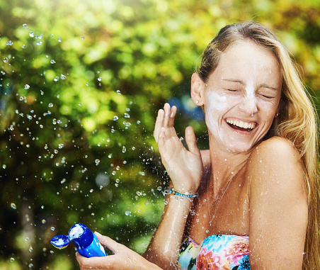Beautiful young woman with her face covered in sunscreen, and holding the container, reacts with laughter as someone out of frame splashes her with drops of water.