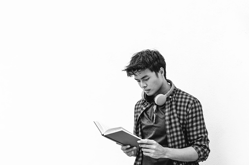 Asian hipster young man reading a book against a white wall - Asian student holding in hands a book and reading this during leisure time outdoors - Leisure activity concept for young people