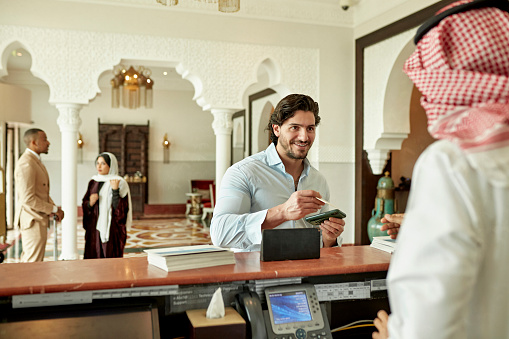 View from beside receptionist in traditional Saudi attire of bearded man holding smart phone and smiling while paying his bill with other guests in background.