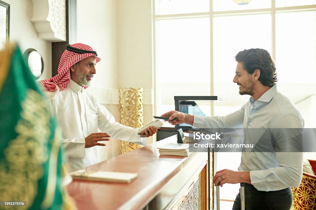 Guest making contactless payment when checking out of hotel Waist-up side view of early 30s Middle Eastern businessman and late 40s male receptionist managing bill at front desk of small luxury hotel in Riyadh. Saudi Arabia Stock Photo