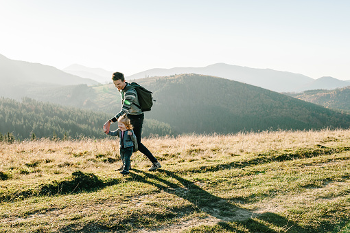 Dad with backpack and child walk in the autumn grass. The daughter and father walking on nature. Family spending time together in mountain on vacation. Holiday trip concept. World Tourism Day.