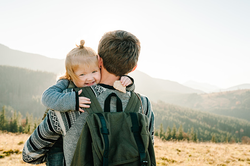 The daughter hug parent on nature. Dad with backpack and child walk in the autumn grass. Family spending time together in mountain outside, on vacation. Holiday trip concept. World Tourism Day.