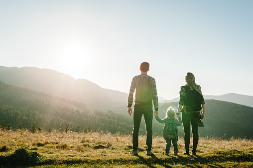 Family with kid hiking in mountains. Young tourists on top of a mountain enjoying valley view sunset. Happy mom, dad with a backpack and daughter. Holiday trip concept. World Tourism Day. Back view.