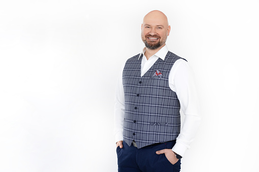 Portrait of smiling happy bald bearded middle-aged man businessman wearing grey checkered vest, white shirt, blue jeans, standing, thrusting hands into pockets, posing on white background. Studio.