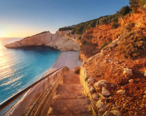 Beautiful stairs on sandy beach at sunset. Porto Katsiki, Lefkada island, Greece. Colorful landscape with blue sea, beautiful rocks in water, steps, empty beach, stones, green trees, sky, sunlight