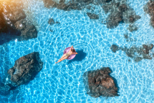 вид с воздуха на молодую женщину, плавающую с розовым кольцом в синем море на закате летом. тропический пейзаж с девушкой, камнями и скалами  - sea swimming greece women стоковые фото и изображения