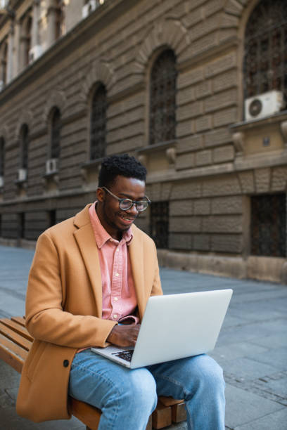 istock Young black student sitting on a bench outside, studying on his laptop with a smile on his face 1392419757