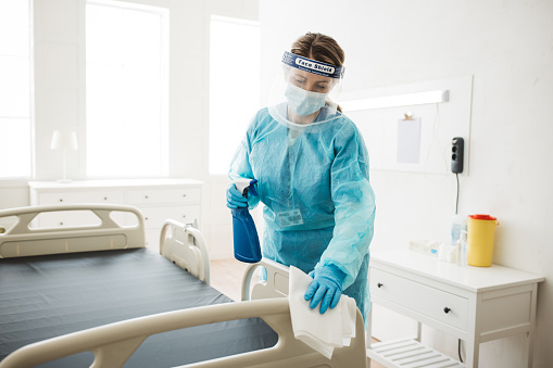 Professional nurse wiping the bed and cleaning room after patients in hospital ward. She is wearing face mask and protective uniform.