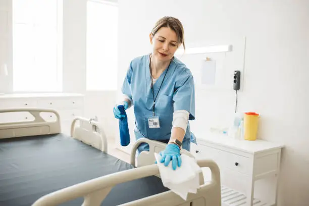 Professional nurse wiping the bed and cleaning room after patients in hospital ward.