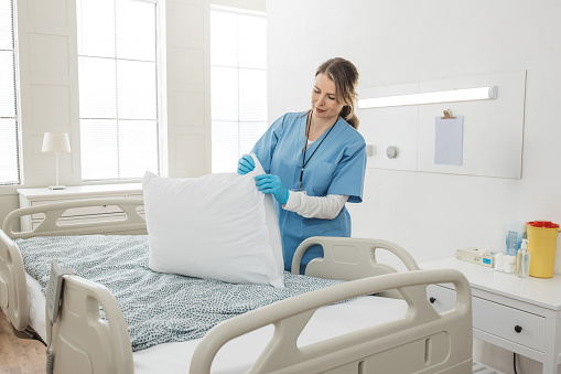 Professional nurse changing sheets on the bed and cleaning room after patients in hospital ward.
