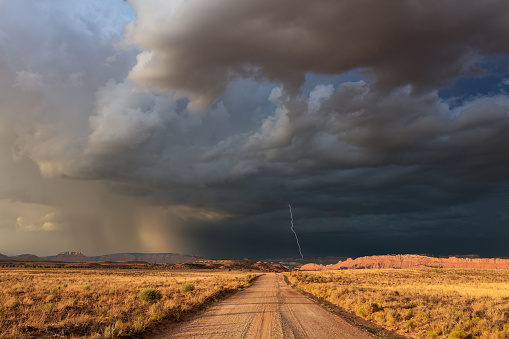 A dirt road leading to ominous, dark storm clouds and a lightning strike over a desert landscape in Utah.