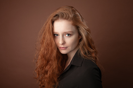 Close-up studio portrait of an attractive 20 year old red-haired woman in a black blouse on a brown background