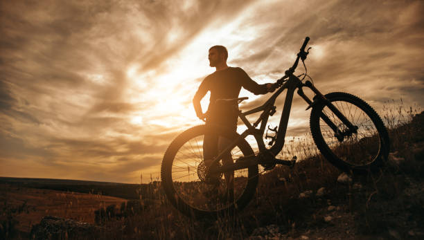 istock Man with bicycle standing on hill at sunset 1392429024