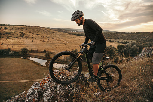 Side view of man in helmet standing on mountain bike on edge of cliff in evening in countryside