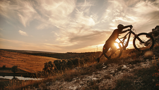 Side view of unrecognizable sportive cyclist climbing up hill with electric mountain bike against sunset sky in summer evening in countryside
