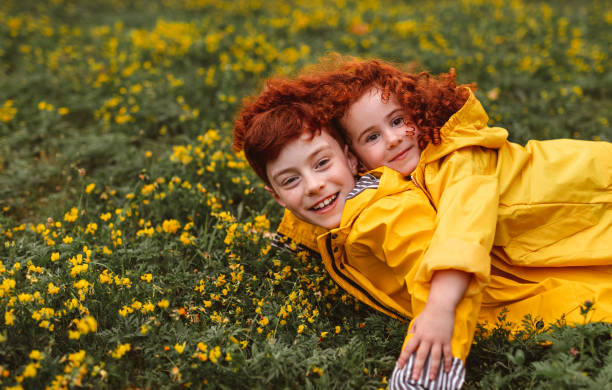Side view of cheerful ginger brother and sister wearing vivid yellow raincoats lying on blooming flower field and having fun
