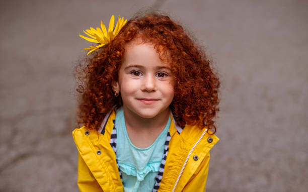 Cute ginger haired girl with floral accessory Adorable little girl with red curly hair wearing bright raincoat with flower on head looking at camera with gentle smile little redhead girl stock pictures, royalty-free photos & images