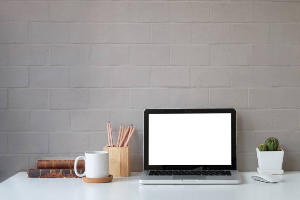istock Front view computer laptop with blank screen, coffee cup, pencil holder and book on white table with brick wall. 1392446518