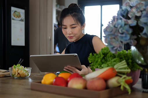 Woman sitting at kitchen table full of fresh vegetables and searching recipes on digital tablet.