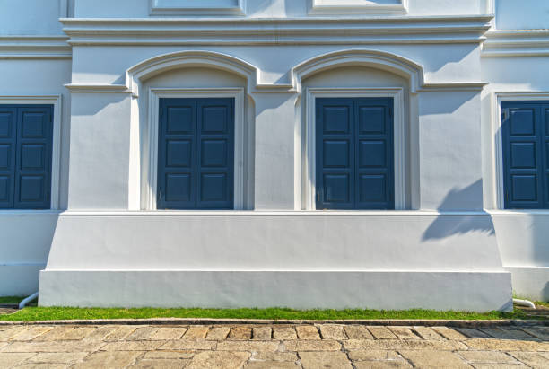 Beautiful background of the white wall of a vintage building and cobblestone street in the front, vintage closed windows, light and shadow from sunset.
