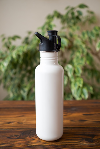 White stainless steel reusable water bottle in on brown rustic wooden table against green foliage background.