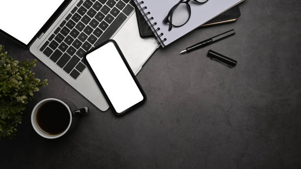 istock Mobile phone with blank screen, laptop computer, coffee cup and eyeglasses on dark stone background. Top view. 1392472250