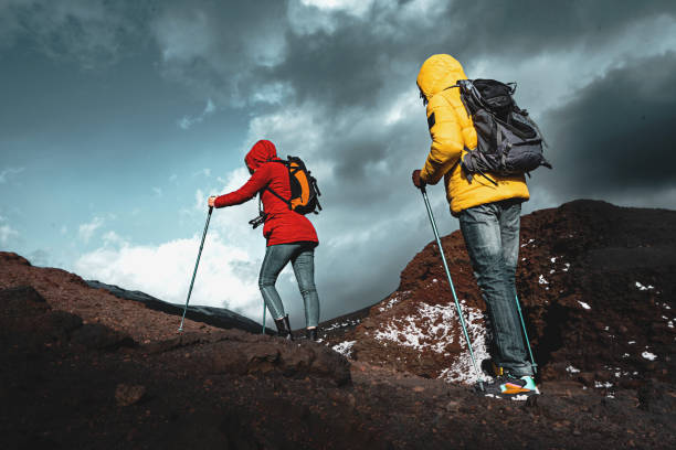 couple of active young people hikers with backpack and trekking poles climbing on the mount etna in sicily, italy - adventure travel and wanderlust concept - mt etna imagens e fotografias de stock