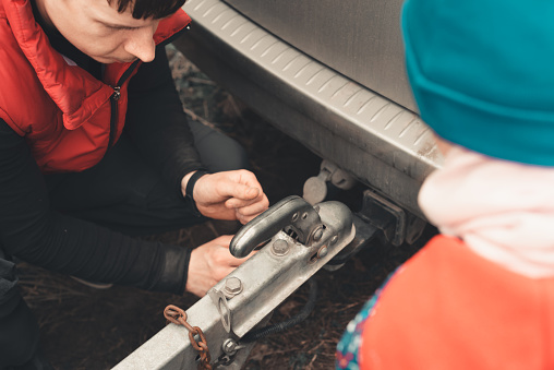 Man connects a trailer to the towbar of his car, and a child watches the process. Travel, cargo transportation or garbage collection concept