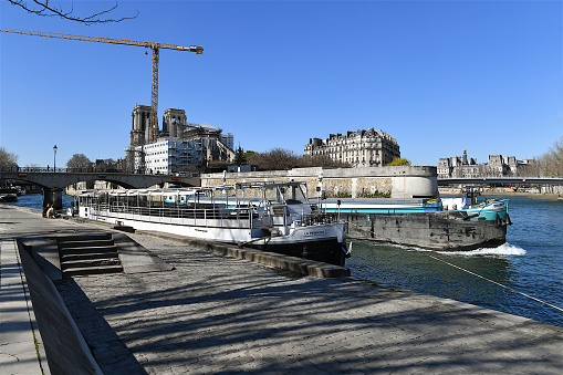 Paris, France-03 18 2022: Moored Houseboat and passing barge  on the Seine river nearby the Notre-Dame Cathedral under rebuilding, Paris, France.