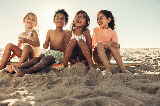 Feeling blissful at the beach. Adorable little kids laughing cheerfully while sitting together on beach sand. Group of carefree young friends having fun together during summer vacation.