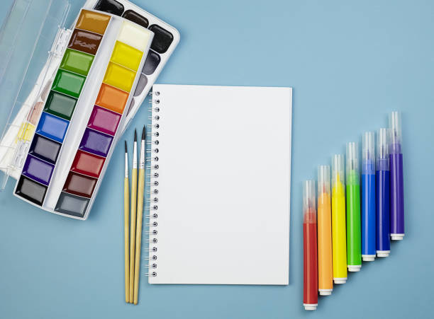 istock Watercolor paints, brushes, an empty spring notebook and multi-colored markers on a blue background with copy space. 1392489966