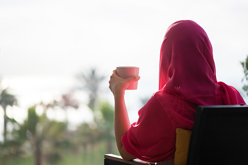 Silhouette of muslim, arab woman in red head scarf, hijab sitting on the chair on terrace with cup of coffee opposite sunrise palm sea beach