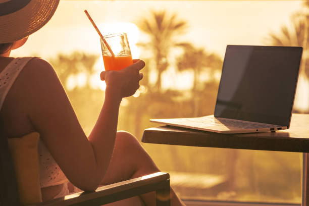 Silhouette of woman freelancer in straw hat drinking juice during working on laptop sitting on the armchair near table on balcony opposite sunset palm sea beach. Remote work on the vacation. Rear view silueta de una mujer freelancer con sombrero de paja bebiendo jugo durante el trabajo en la computadora portátil sentada en el sillón cerca de la mesa en el balcón frente a la playa de mar de palmeras al atardecer. trabajo remoto en las vacaciones. vist
