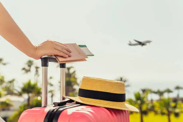 Woman with pink suitcase and passport with boarding pass standing on passengers ladder of airplane opposite sea with palm trees. Tourism concept