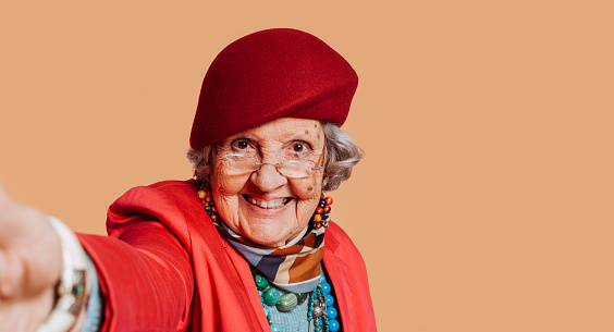 Cheerful stylish elderly grandmother taking a selfie. Old female wears red beret, pink suit and some accessories. Portrait at studio. Senior old woman looking at camera. Copy space.