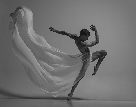 Black and white portrait of graceful muscled male ballet dancer dancing with fabric, cloth isolated on grey studio background. Grace, art, beauty, contemp dance concept. Weightless, flexible actor