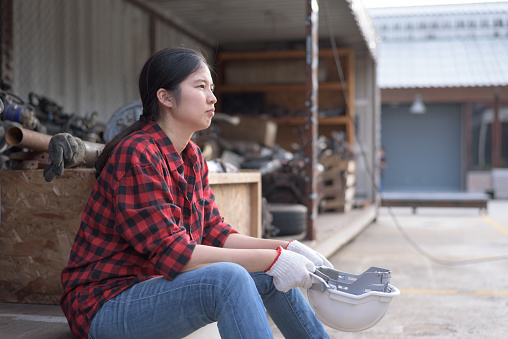 Side view portrait of an Asian woman in red lumberjack shirt holding safety hardhat, sitting, resting, smiling, relaxing, looking away. Female worker, blue-collar labor, foreman or engineer concept