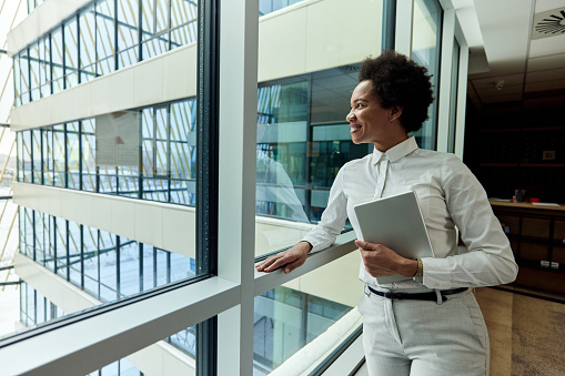 Happy African American businesswoman holding touchpad and day dreaming while looking through a window from a hallway of an office building.