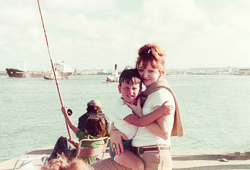 Vintage image of a mother holding her son and looking at the camera at a fishing deck with fisherman and fisher boats in the background. Vintage image from the seventies.