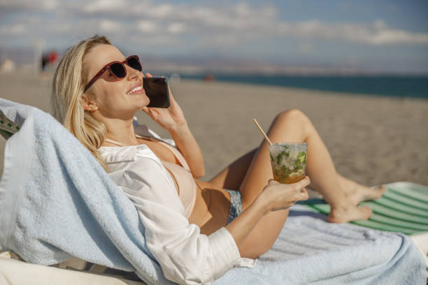 istock Smiling lady lying on deckchair, having call and drinking cocktail on a beach 1392513916