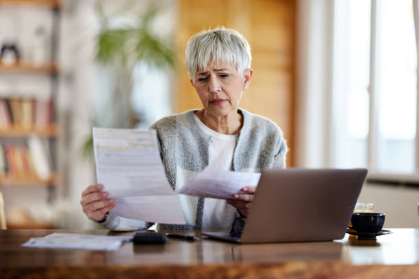 Worried senior woman paying her bills over laptop at home. Mature woman feeling worried while going through her bills at home. cost of living stock pictures, royalty-free photos & images