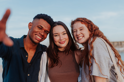 Three diverse friends smiling at the camera while standing next to the sea outdoors. Group of happy friends having a good time while on a weekend getaway in a coastal town.