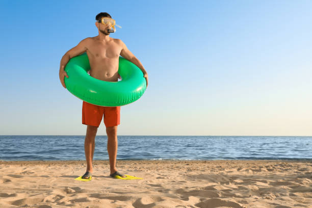 istock Man with flippers, inflatable ring and goggles on sea beach. Space for text 1392520670