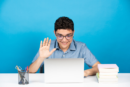 Hispanic young man greeting to webcam in a video call. Teenager boy in video conference in front of laptop isolated on blue background