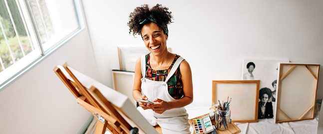 Cheerful female painter holding a smartphone in her art studio. Happy young female painter smiling at the camera while sitting in front of her canvas. Young woman updating her painting blog online.